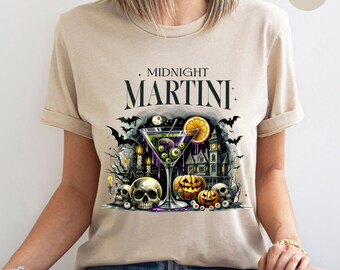 May include: Sand-colored t-shirt featuring a Halloween-themed graphic. The design includes a martini glass filled with green liquid and eyeballs, a haunted house, pumpkins, skulls, and the words "MIDNIGHT MARTINI". Bats and candles complete the spooky scene.