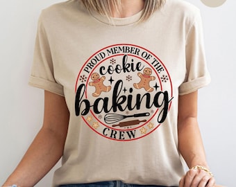 Peut inclure: Un t-shirt couleur sable avec un motif circulaire portant l'inscription "PROUD MEMBER OF THE COOKIE BAKING CREW". Le motif comprend des bonhommes en pain d'épices, des étoiles, un fouet et un rouleau à pâtisserie. Le t-shirt a des manches courtes et un col rond.