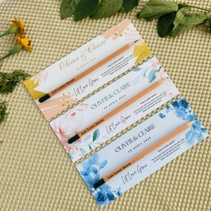 May include: Three wedding favor cards with a floral design and the text "Let Love Grow" printed on them. Each card has a wooden pencil with a black eraser attached to it. The cards are decorated with watercolor flowers in pink, blue, and green. The text on the cards reads "Oliver & Claire" and "20 April 2024".