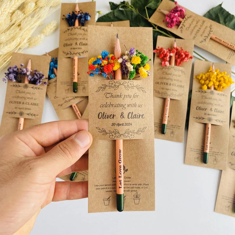 May include: A collection of pencils with colorful dried flowers attached to kraft paper cards. The cards read "Thank you for celebrating with us Oliver & Claire 20 April 2024" and "Let Love Grow".