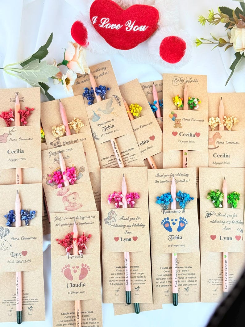 Puede incluir: Una colecci&oacute;n de recuerdos de fiesta personalizados con l&aacute;pices con detalles florales y tarjetas impresas. Las tarjetas incluyen nombres, fechas y frases como "Thank you for celebrating my birthday" y "Prima Comunione". Un coraz&oacute;n rojo con "I Love You" est&aacute; en el fondo.