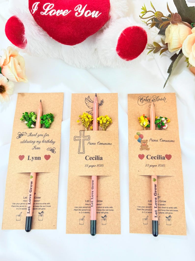 Puede incluir: Tres l&aacute;pices personalizados con sobres de semillas. Cada l&aacute;piz se muestra en una tarjeta de papel kraft con texto y elementos decorativos. Uno dice "Gracias por celebrar mi cumplea&ntilde;os de Lynn". Los otros dos dicen "Prima Comunione Cecilia 22 giugno 2025" y "Baby Shower Cecilia 22 giugno 2025."