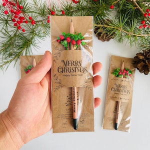 以下が含まれることがあります： クリスマスをテーマにした鉛筆。茶色の紙に「Merry Christmas」の文字が印刷され、上部に花の装飾が施されています。鉛筆の先端は黒で、緑と赤い実で飾られています。