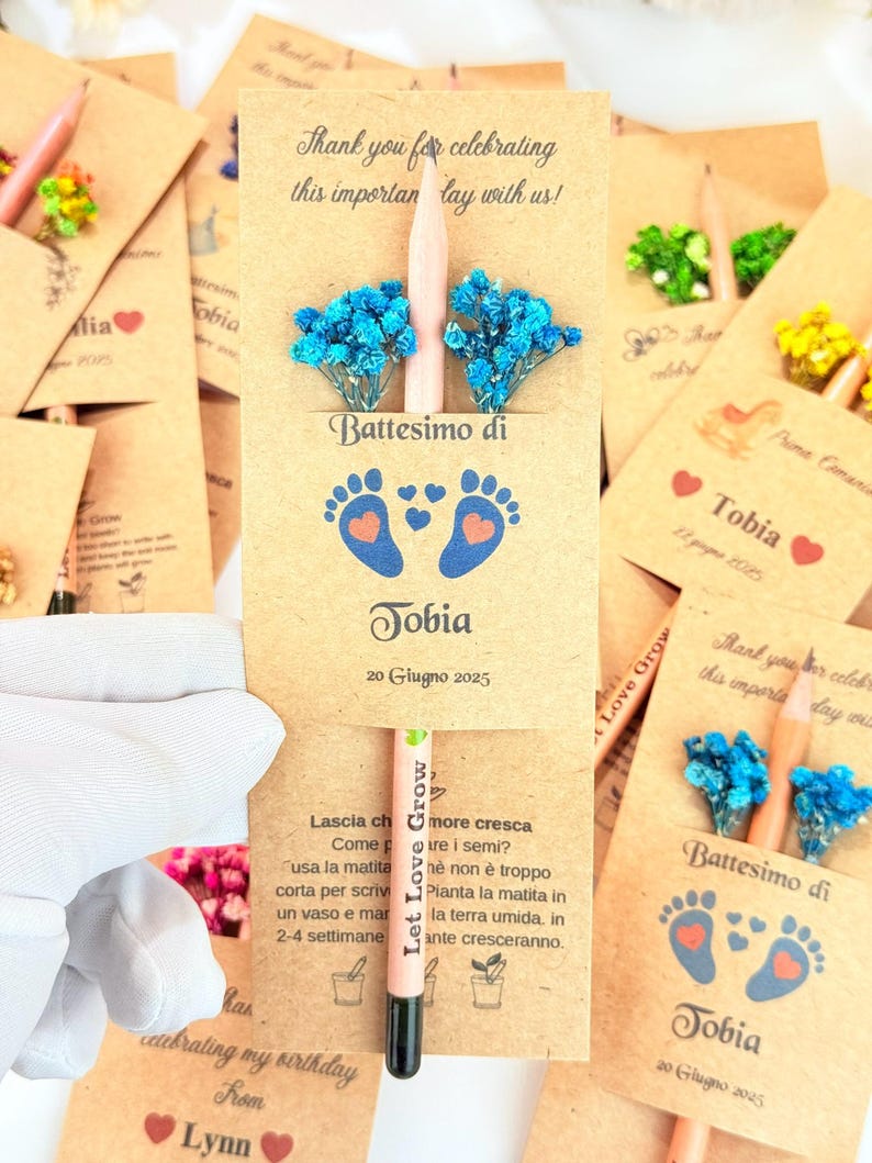 Puede incluir: Recuerdos de papel kraft para un bautizo, cada uno con un l&aacute;piz, flores azules y el nombre "Tobia" y la fecha "20 Giugno 2025". Tambi&eacute;n se ve el texto "Thank you for celebrating this important day with us!"