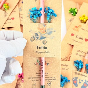 Puede incluir: Un l&aacute;piz rosa con el texto "Let Love Grow" se muestra en un recuerdo de baby shower. El recuerdo incluye una tarjeta con una ilustraci&oacute;n de oso de peluche en un avi&oacute;n, flores azules y el nombre "Tobia" con una fecha.