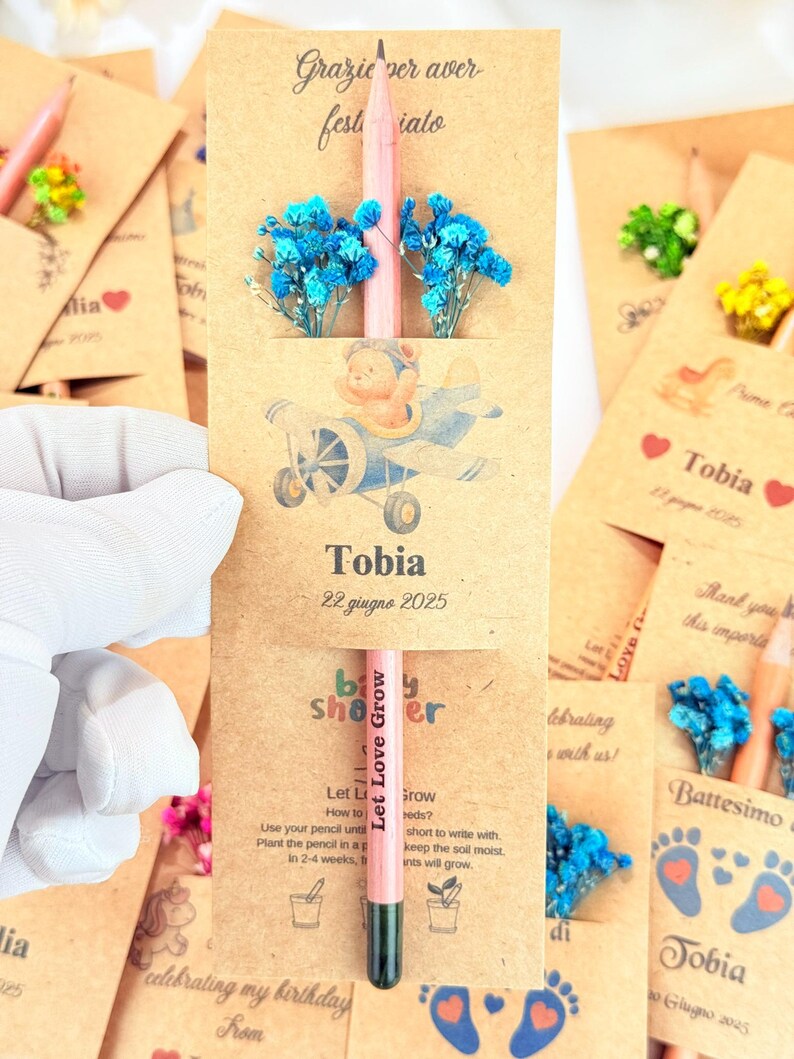 Puede incluir: Un l&aacute;piz rosa con el texto "Let Love Grow" se muestra en un recuerdo de baby shower. El recuerdo incluye una tarjeta con una ilustraci&oacute;n de oso de peluche en un avi&oacute;n, flores azules y el nombre "Tobia" con una fecha.