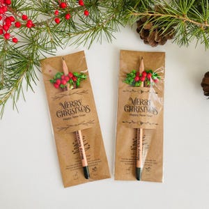 以下が含まれることがあります： 「MERRY CHRISTMAS Happy New Year」の文字が入った茶色の紙パッケージに入った2本の鉛筆。鉛筆は黒い先端とクリスマスツリーのデザインです。パッケージは赤いベリーと緑の葉で飾られています。