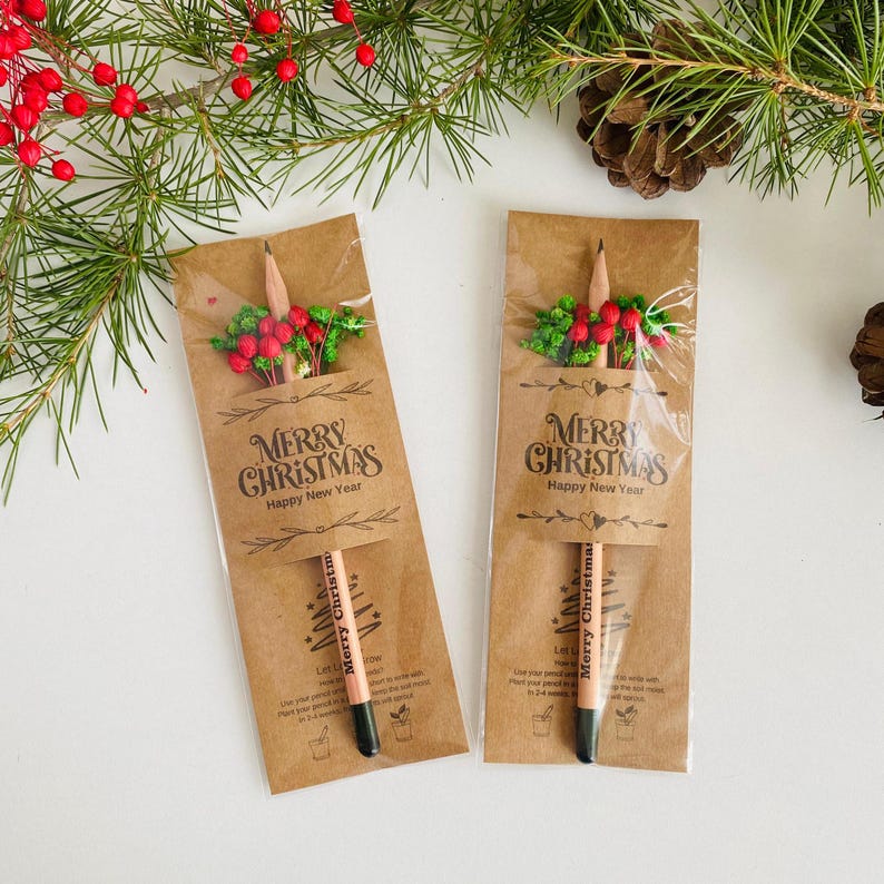 以下が含まれることがあります： 「MERRY CHRISTMAS Happy New Year」の文字が入った茶色の紙パッケージに入った2本の鉛筆。鉛筆は黒い先端とクリスマスツリーのデザインです。パッケージは赤いベリーと緑の葉で飾られています。