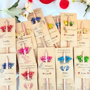 Puede incluir: Una colecci&oacute;n de l&aacute;pices personalizados para eventos especiales. Cada l&aacute;piz se combina con un arreglo floral decorativo y una tarjeta con texto personalizado. Las tarjetas presentan nombres, fechas y detalles del evento, con una almohada roja en forma de coraz&oacute;n en el fondo.