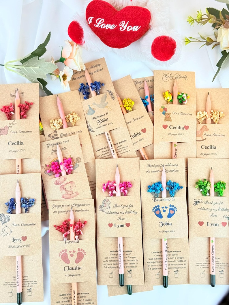 Puede incluir: Una colecci&oacute;n de l&aacute;pices personalizados para eventos especiales. Cada l&aacute;piz se combina con un arreglo floral decorativo y una tarjeta con texto personalizado. Las tarjetas presentan nombres, fechas y detalles del evento, con una almohada roja en forma de coraz&oacute;n en el fondo.