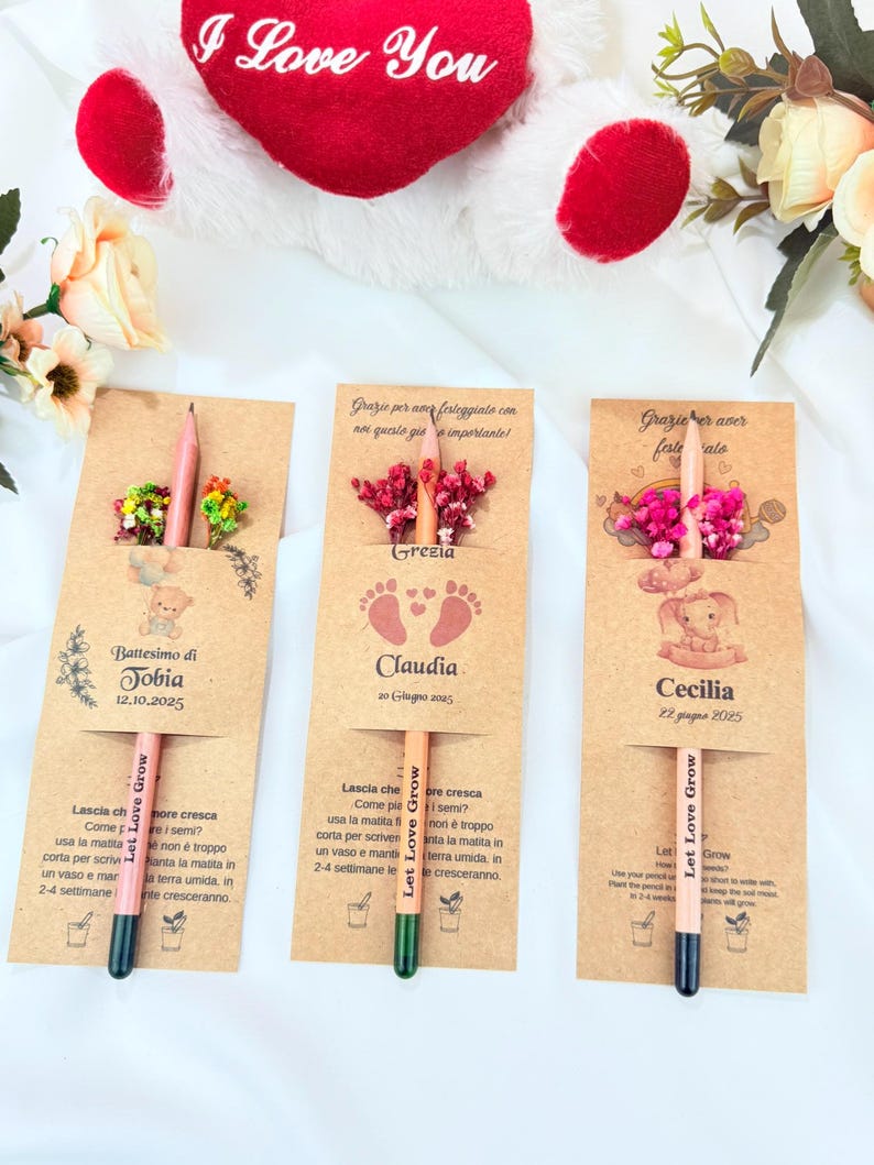 Puede incluir: Tres recuerdos de l&aacute;pices personalizados con detalles florales. Cada l&aacute;piz est&aacute; unido a una tarjeta con un nombre y una fecha, con el texto "Let Love Grow". Un peluche rojo en forma de coraz&oacute;n con "I Love You" est&aacute; en el fondo.