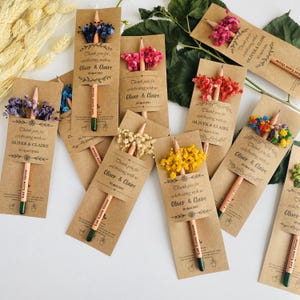 May include: A collection of kraft paper favor cards with pencils and dried flowers. Each card reads "Thank you for celebrating with us Oliver & Claire 30 April 2024" and "Let Love Grow". The pencils have green tips and are decorated with various colored flowers.