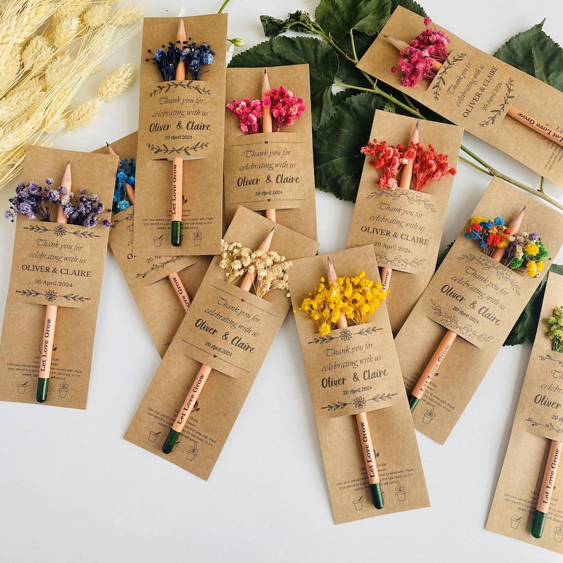 May include: A collection of kraft paper favor cards with pencils and dried flowers. Each card reads "Thank you for celebrating with us Oliver & Claire 30 April 2024" and "Let Love Grow". The pencils have green tips and are decorated with various colored flowers.