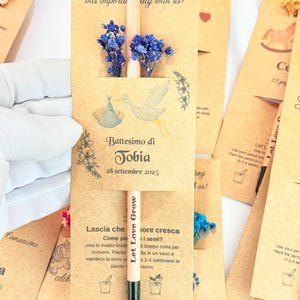 Puede incluir: Recuerdos de bautizo de papel kraft, con un l&aacute;piz con la frase "Let Love Grow" y flores azules secas. El dise&ntilde;o incluye una ilustraci&oacute;n de cig&uuml;e&ntilde;a y el texto "Battesimo di Tobia 28 Settembre 2025".