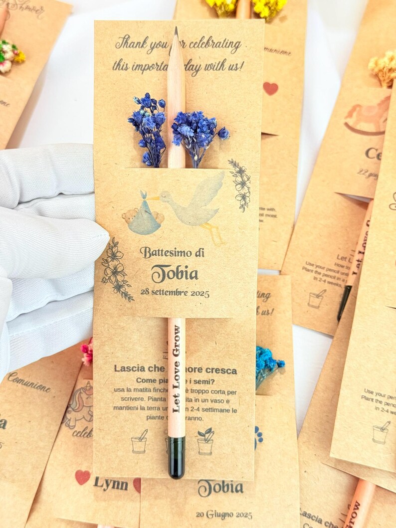 Puede incluir: Recuerdos de bautizo de papel kraft, con un l&aacute;piz con la frase "Let Love Grow" y flores azules secas. El dise&ntilde;o incluye una ilustraci&oacute;n de cig&uuml;e&ntilde;a y el texto "Battesimo di Tobia 28 Settembre 2025".