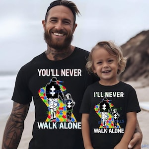 May include: Black t-shirts with the text "YOU'LL NEVER WALK ALONE" and "I'LL NEVER WALK ALONE". Each shirt features a graphic of a person and child walking together, with a puzzle piece design.