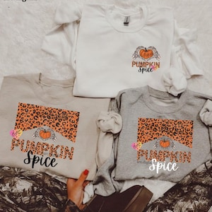 May include: Three sweatshirts in white, tan, and gray, each featuring a pumpkin spice design. The design includes a leopard print background, a pumpkin, and the words "Pumpkin Spice". The sweatshirts are casual wear.