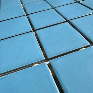 May include: A close-up shot of a grid of square, light blue tiles. The tiles have a glossy finish and are arranged in neat rows. The grout lines between the tiles are dark, creating a contrast with the bright blue color.