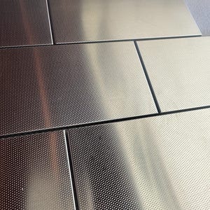 May include: Close-up of several rectangular, metallic tiles. The tiles have a shiny, silver finish and are arranged in a grid pattern. Each tile is covered in a pattern of tiny, evenly spaced perforations, creating a textured surface.