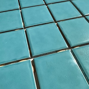 May include: A close-up shot of a grid of square, turquoise-colored tiles. The tiles have a glossy finish and are arranged closely together, with thin, dark grout lines separating them. The overall effect is a smooth, uniform surface.