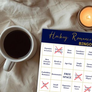 Puede incluir: Una tarjeta de juego "Hockey Romance Bingo" azul marino y blanca con una taza de caf&eacute; y una vela encendida. La tarjeta de bingo presenta tropos de novelas rom&aacute;nticas, con algunas casillas marcadas con una X.