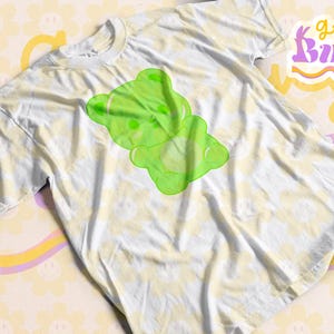 Puede incluir: Una camiseta blanca con un gr&aacute;fico de un oso de gominola verde. La camiseta tiene un patr&oacute;n de tie-dye y el texto "groovy bunny co." es visible en el fondo.