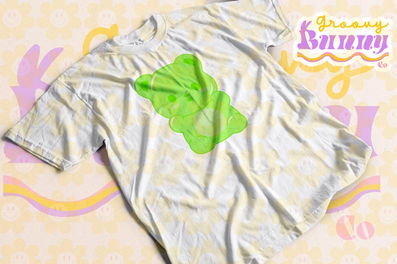 Puede incluir: Una camiseta blanca con un gr&aacute;fico de un oso de gominola verde. La camiseta tiene un patr&oacute;n de tie-dye y el texto "groovy bunny co." es visible en el fondo.