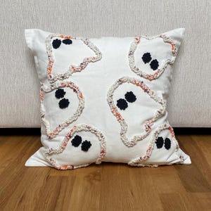 May include: White square decorative pillow featuring embroidered ghost designs with black eyes and colorful trim. The ghosts are outlined with a textured, multi-colored yarn. A festive accent for Halloween or spooky decor.