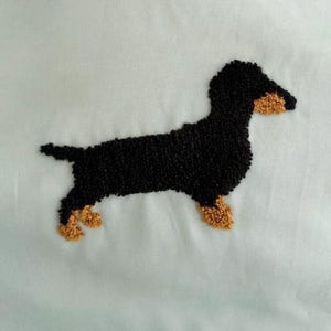 May include: Embroidered dachshund dog in black and tan. The dog is depicted in a side profile, with its body and head in black and its paws and snout in tan. The background is a light blue fabric.