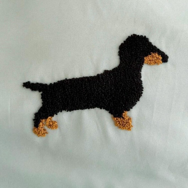 May include: Embroidered dachshund dog in black and tan. The dog is depicted in a side profile, with its body and head in black and its paws and snout in tan. The background is a light blue fabric.
