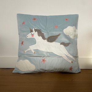 Op de afbeelding: Een lichtblauw vierkant kussen met een wit eenhoornontwerp. De eenhoorn heeft grijs haar en een grijze vleugel, tegen een lichtblauwe achtergrond met geborduurde witte wolken en roze sterren. Het kussen is gemaakt van zachte stof.