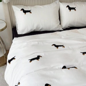 May include: White bedding set featuring embroidered black dachshund dogs. The set includes two pillowcases and a duvet cover, creating a cohesive and charming aesthetic. The dachshunds are detailed with brown accents, adding a touch of warmth to the design.