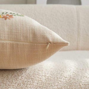 May include: Close-up of a beige decorative pillow with embroidered floral details in yellow, pink, and green. The pillow has a zipper closure and is made of a textured fabric. The pillow is on a textured surface.
