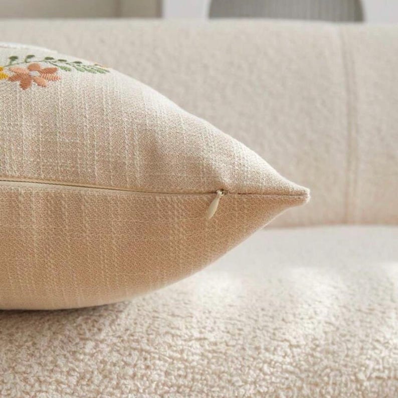 May include: Close-up of a beige decorative pillow with embroidered floral details in yellow, pink, and green. The pillow has a zipper closure and is made of a textured fabric. The pillow is on a textured surface.