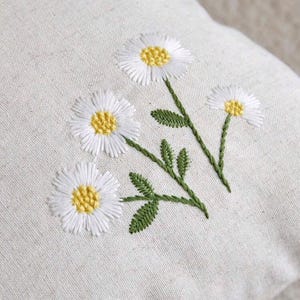 May include: A light beige fabric cushion cover featuring an embroidered design of three white daisy flowers with yellow centers and green stems with small leaves. The embroidery is stitched directly onto the textured, woven natural-colored textile.