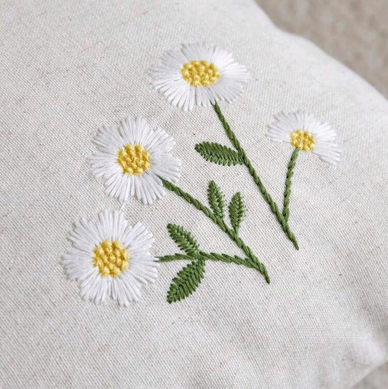 May include: A light beige fabric cushion cover featuring an embroidered design of three white daisy flowers with yellow centers and green stems with small leaves. The embroidery is stitched directly onto the textured, woven natural-colored textile.