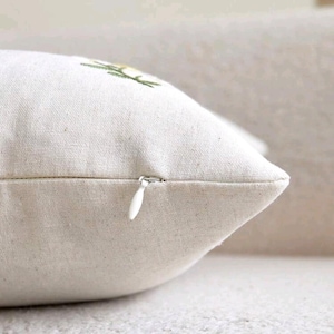 May include: A square throw pillow cover made of off-white, textured linen-like fabric. The cover features a hidden white zipper closure along the side seam and a small portion of a green and yellow embroidered floral design visible at the top.