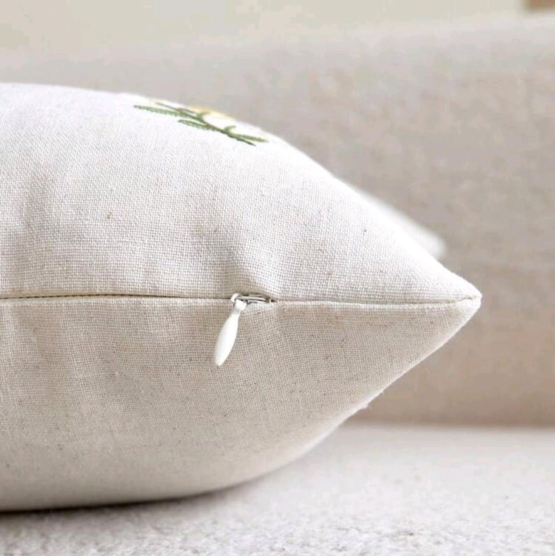 May include: A square throw pillow cover made of off-white, textured linen-like fabric. The cover features a hidden white zipper closure along the side seam and a small portion of a green and yellow embroidered floral design visible at the top.