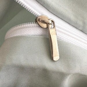 May include: Close-up of a light green fabric with a white zipper. The zipper pull is a gold color. The fabric has a subtle texture.