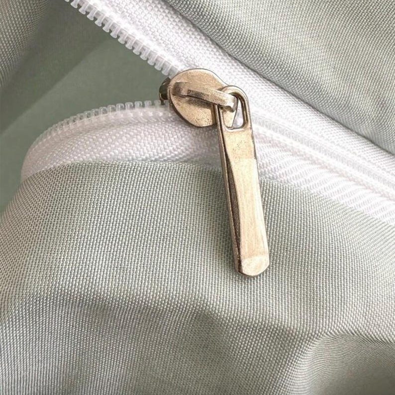 May include: Close-up of a light green fabric with a white zipper. The zipper pull is a gold color. The fabric has a subtle texture.