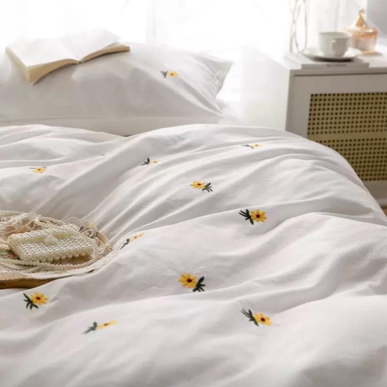 May include: White bedding set with embroidered yellow flower details. The set includes a pillowcase and a duvet cover, with a book on the pillow. A tray with a decorative item is in the foreground. The bedding is made of a soft, possibly linen, material.