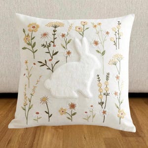 May include: Square white decorative pillow featuring a white felt bunny silhouette and embroidered floral designs in yellow, green, and white. The pillow has a soft, textured appearance, perfect for adding a touch of spring to any room.