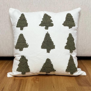 May include: White square throw pillow with nine olive green textured Christmas tree designs. The pillow is made of a soft fabric and is placed on a wooden floor. The pillow is a decorative item.