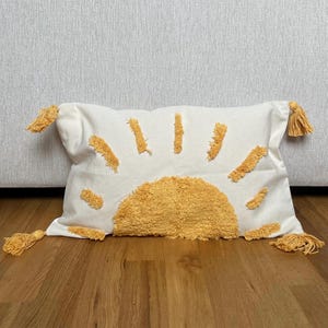 May include: A white rectangular decorative pillow with a yellow sunburst design. The sun is made of textured fabric, and the rays are embroidered. The pillow has yellow tassels on each corner.