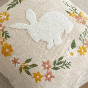 May include: Cream-colored square pillow with a white, textured bunny applique in the center. The pillow is embroidered with a floral wreath in yellow, pink, and green, creating a spring or Easter theme.