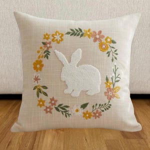 May include: Square decorative pillow with a cream-colored background. The pillow features a white bunny in the center, surrounded by a floral wreath in yellow, pink, and green. The pillow is likely made of linen or a similar fabric.