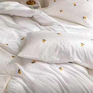 May include: White bedding set featuring embroidered yellow floral designs. The set includes a duvet cover and pillowcases. The fabric appears soft and inviting, perfect for a bedroom refresh. The floral pattern adds a touch of spring to the bedroom.
