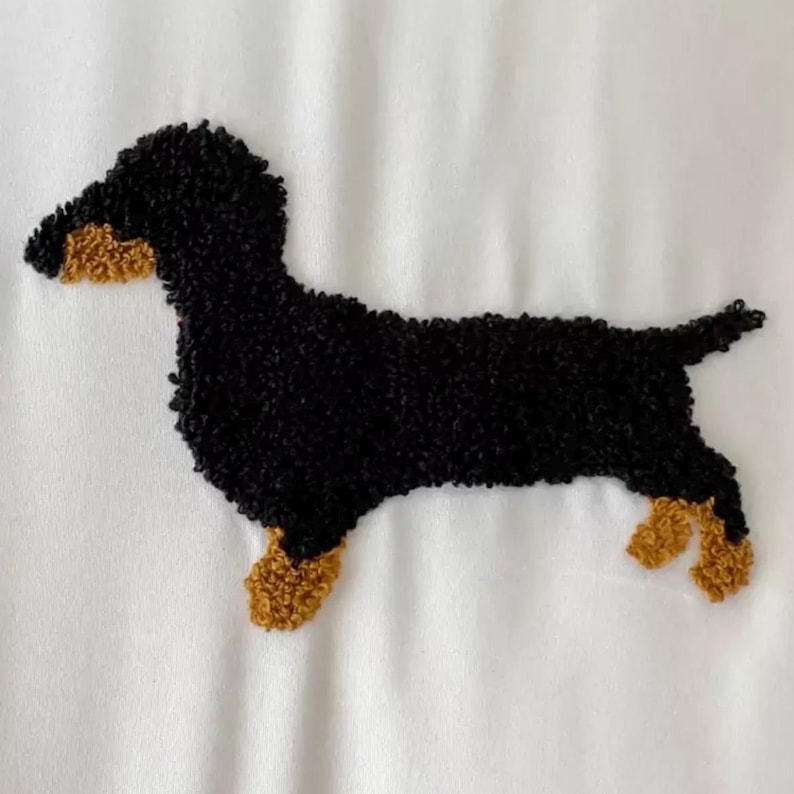May include: A black and tan dachshund dog design on a white background. The dog is depicted in a side profile, with its body and head in black and its paws and snout in tan. The design has a textured, embroidered appearance.