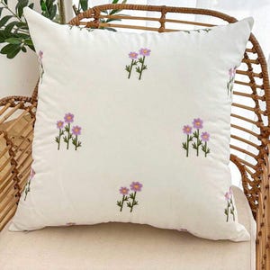 May include: A square, off-white pillow with embroidered purple flowers and green stems. The pillow is placed on a wicker chair, showcasing its decorative floral design. The pillow's soft texture and floral pattern add a touch of elegance to any room.