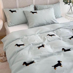 May include: A light blue bedding set featuring embroidered black dachshund dogs. The set includes a duvet cover and pillowcases. The bed is made up with a white mattress and a woven headboard.